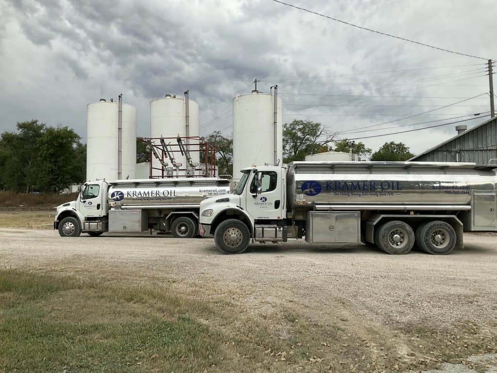 Two fuel delivery trucks labeled "Kramer Oil" are parked in front of large white storage tanks on a gravel lot. The sky is overcast, and a metal building stands in the background, underscoring the efficiency championed by Chad Kramer as President of Fuel True.